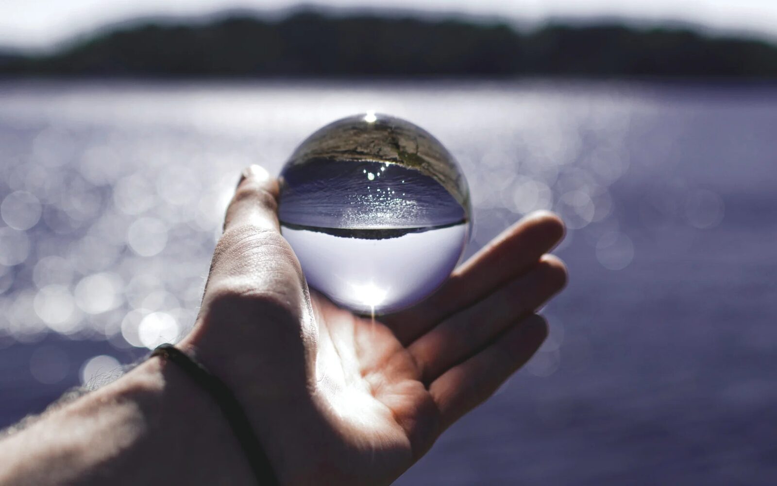 An open hand holding a glass sphere in front of a sunlit lake